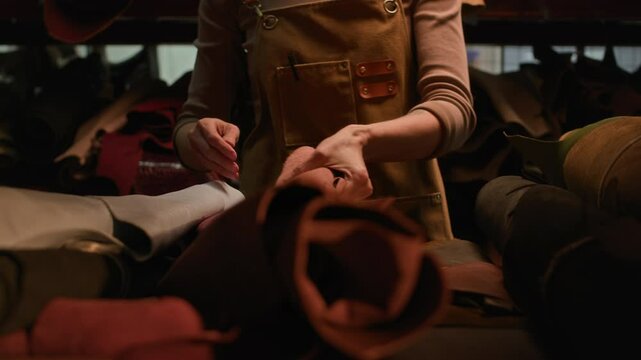 Midsection shot of unrecognizable female artisan wearing apron sorting out leather rolls in dim lit tannery storehouse and taking one away