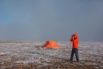 Man in red jacket in hood with photo camera near orange tent on snow-covered stony pass in sunlight...