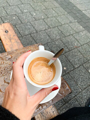 Close-up of a hand holding a cup of espresso with foam, wooden bench and wet pavement visible.