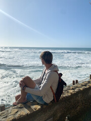 Man sitting on a stone wall overlooking ocean waves under clear blue sky.