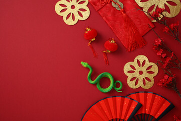 Chinese New Year decorations on a red background, featuring gold floral cutouts, red lanterns, a green snake, red fans, and cherry blossoms