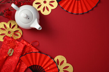 Chinese New Year celebration background with a white teapot, red fans, golden decorations, and red envelopes on a red surface