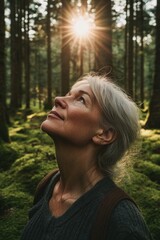 A mature woman with grey hair stands in a misty forest, looking upwards towards the bright sunlight breaking through the trees. She is dressed in casual attire and appears contemplative.