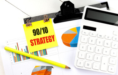 Yellow sticky note with the words 90 10 Strategy on a clipboard, placed over financial charts with...