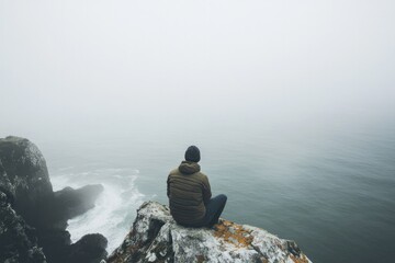 Sitting on rocky edge, figure contemplates misty ocean below, se