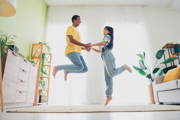 Photo of funny funky positive multinational couple have fun jump up good mood weekend vacation indoors flat apartment