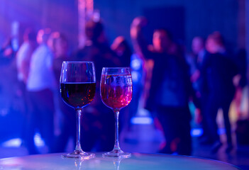 Two wine glasses sit on a table at a party with colorful lighting and blurred dancing crowd in background, capturing festive atmosphere.