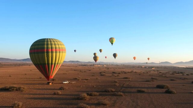 Hot air balloons soar over a desert landscape at sunrise, creating a serene and colorful scene. Clear blue sky and distant mountains enhance the atmosphere.