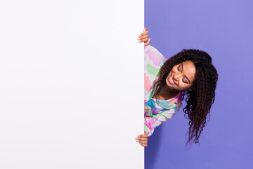 Joyful young woman with curly hair peeking from behind a white panel against a vibrant purple...