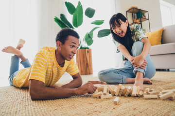 Photo of crazy funny charming positive multinational couple sit floor play jenga tower fall down indoors flat