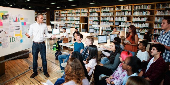 A diverse group of people in a library setting, listening to a presentation. The audience is engaged, with a mix of genders and ethnicities, focused on the speaker. Teacher teaching high school class.