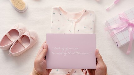 Baby shower gift with pink card, baby shoes, and onesie. Soft pink theme for baby girl. Hands holding card. Baby shower essentials for newborn celebration. Pastel pink girl baby shower, top view image