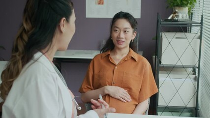 Medium shot of young female doctor consulting Asian woman about her pregnancy during appointment at clinic