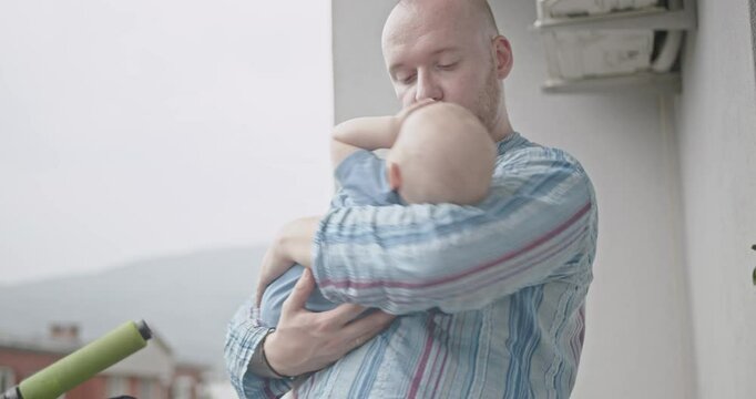 A father holds his baby on the balcony, gently bouncing and kissing the child. The baby looks happy and comfortable in his father's arms.