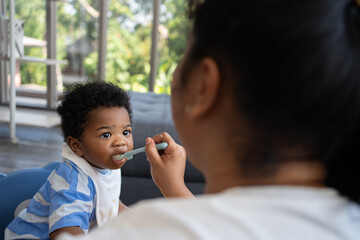 Asian mother feeding her 9 months old her cute little baby and African American helping for holding food plate At Home. Photo series of family, kids and happy people concept. Parents feed kids.