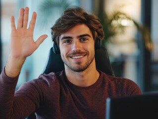 Young man smiling and waving from computer desk, friendly demeanor in professional office setting.