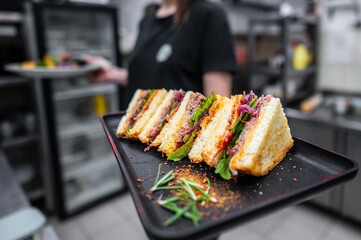 A close-up of a neatly arranged sandwich platter on a black tray, featuring layers of meat, greens, and herbs. A person partially visible in the background, enhancing the culinary setting.