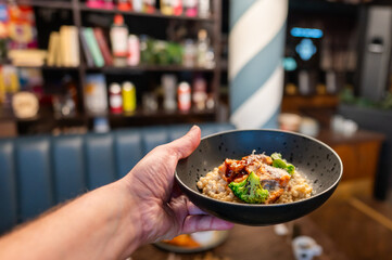 A hand holding a bowl of healthy grain dish topped with broccoli and sauce, set against a stylish cafe background with colorful decorations and shelves. Perfect for food and lifestyle themes.