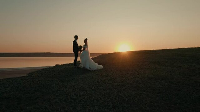 Wedding couple on sunset background. Beautiful bride and groom near the ocean. Honeymoon romantic couple in love holding hands walking in nature. Lovers or newlywed married young couple by the sea.