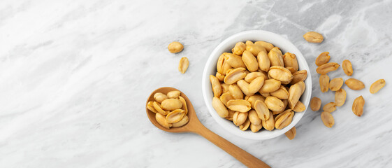 Roasted peanuts in white bowl with wooden spoon on marble table background, top view
