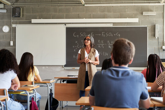 Mature female teacher explaining chemistry formulas and equations written on blackboard to diverse high school students sitting at desks in a classroom. Image with copy space.