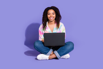 Naklejka premium Young woman with curly black hair and casual attire sitting cross-legged with a laptop against a vibrant purple background