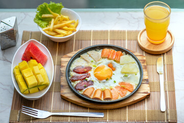 A plate of food with an egg in the center, surrounded by sausage, ham, and other breakfast items. There is also a side of fruit and a glass of juice.