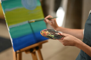 A close-up of an artist hands skillfully blending colors on a palette and adding detailed strokes to a vibrant landscape painting in progress on an easel