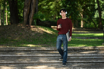 A man wearing headphones holds a coffee cup and a laptop, strolling confidently down outdoor steps...
