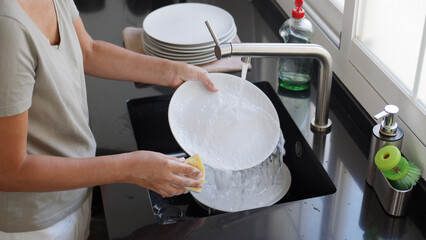Woman washing and rinsing dishes by hand under a stream of water in the kitchen sink. Washing dishes process.