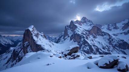 Snow-capped mountains against a deep blue sky dotted with dark gray clouds. Sunlight pierces through, illuminating the peaks in radiant white