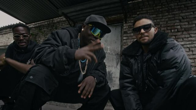 Handheld low angle shot of cool Black male rapper in sunglasses and cap vibing to hip hop music with diverse friends outdoors near abandoned building, they wearing black baggy clothes and chains