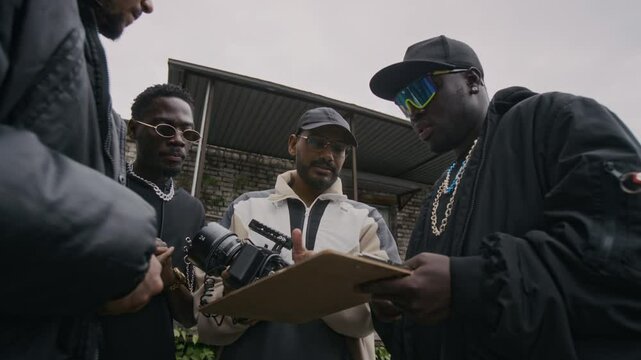 Low angle shot of cool rapper with backup dancers wearing sunglasses, black baggy clothes and chains, discussing camera report with videographer while creating musical video outdoors