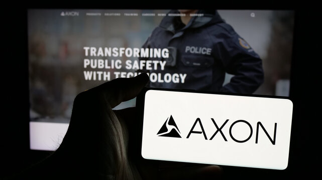 Stuttgart, Germany - 01-16-2025: Person holding mobile phone with logo of US security equipment company Axon Enterprise Inc. on screen in front of web page. Focus on phone display.