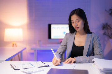 Young asian businesswoman working late at night in office writing notes on documents sitting at desk with computer and paperwork using pen in purple neon light