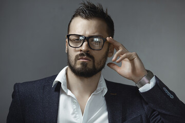 Serious young businessman wearing glasses and a formal suit, striking a thoughtful pose with a hand gesture. Perfect for leadership, corporate, and career-oriented photography themes. 