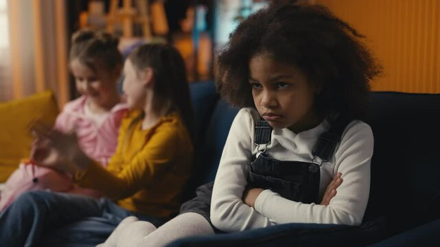 Upset little girl sits alone while her two friends talk nearby, feeling excluded