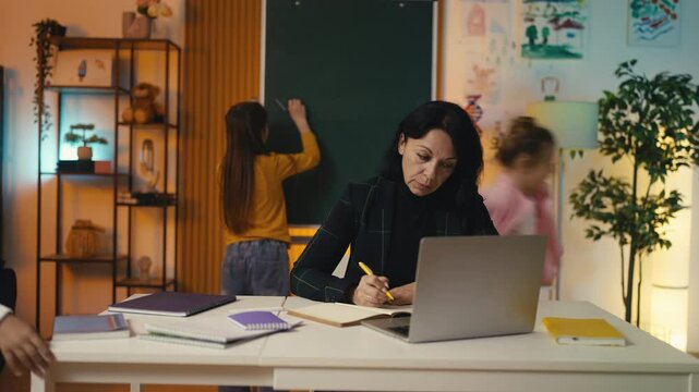 Tired teacher grades student tests while noisy pupils run around the classroom