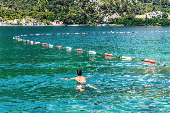 A man swims in the calm waters of Boka Kotorska Bay, Montenegro, surrounded by a scenic mountain landscape. The clear, refreshing water and serene environment make it a perfect spot for relaxation