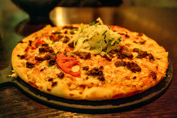 A delicious gourmet pizza topped with fresh tomato slices, melted cheese, savory meat chunks, and a garnish of fresh greens. The warm lighting creates a cozy and appetizing atmosphere