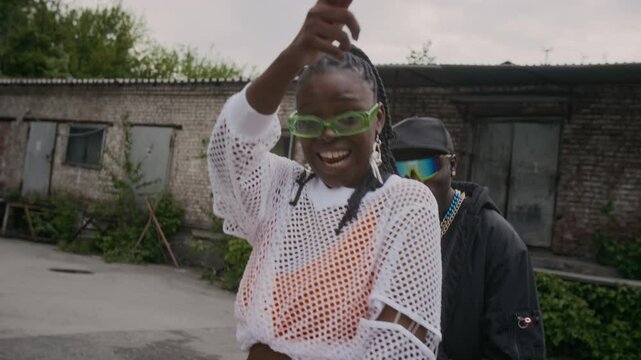 Tilt shot of classy African American lady in glasses and casual clothes singing and vibing to upbeat music with her cool-looking boyfriend while filming music video outdoors in shabby area