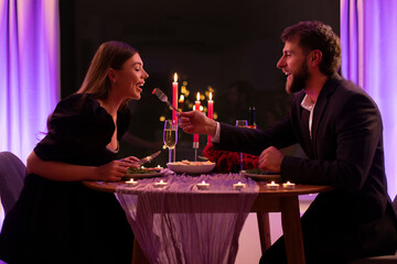 Caring man feeding beloved girlfriend with salad, couple having romantic date in restaurant in the evening, side view shot. Valentine's date