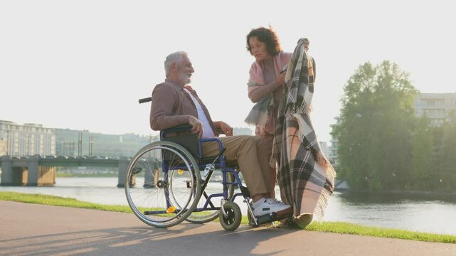 Old man in wheelchair walking with caregiver senior woman in park. Elderly family couple woman supporting taking care of paralyzed man in chair for people with disability outdoor. Rehabilitation