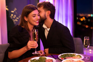 Loving couple dining in restaurant, celebrating Valentine's day together, man whispering into girlfriend's ear and lady smiling