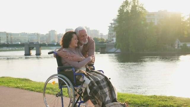 Senior woman in wheelchair walking with caregiver old man on road in park. Elderly family couple man supporting embracing paralyzed woman in chair for people with disability outdoor. Rehabilitation