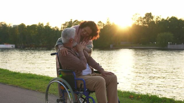 Old man in wheelchair walking with caregiver senior woman on road in park. Elderly family couple woman supporting embracing paralyzed man in chair for people with disability outdoor. Rehabilitation