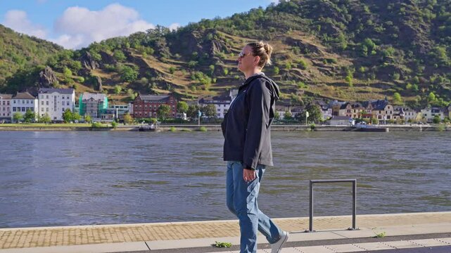 Woman walking along the banks of the Rhine River in the town of Sankt Goar, with the town of St. Goarshausen in the distance, Rhine River Valley, Germany