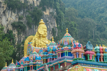 Grandeur of the Batu Caves in Kuala Lumpur, Malaysia. Towering golden statue of Lord Murugan against backdrop of lush green foliage and imposing cave walls. Vibrant colors of Hindu temple complex