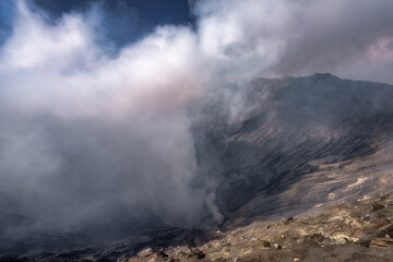 A breathtaking and dramatic volcanic scene is presented, showcasing thick smoke and ash rising gracefully from the Bromo crater against a stunning rugged mountainous backdrop, East Java, Indonesia