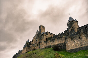 Panoramic view of Carcassonne Castle with medieval stone towers and walls seen from below on a hillside with wet green grass under a cloudy grey sky on a humid day.